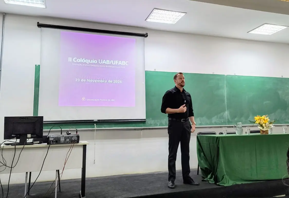 Eduardo Henrique Gomes durante apresentação no II Colóquio UAB/UFABC, em sala de aula com projetor e quadro ao fundo.